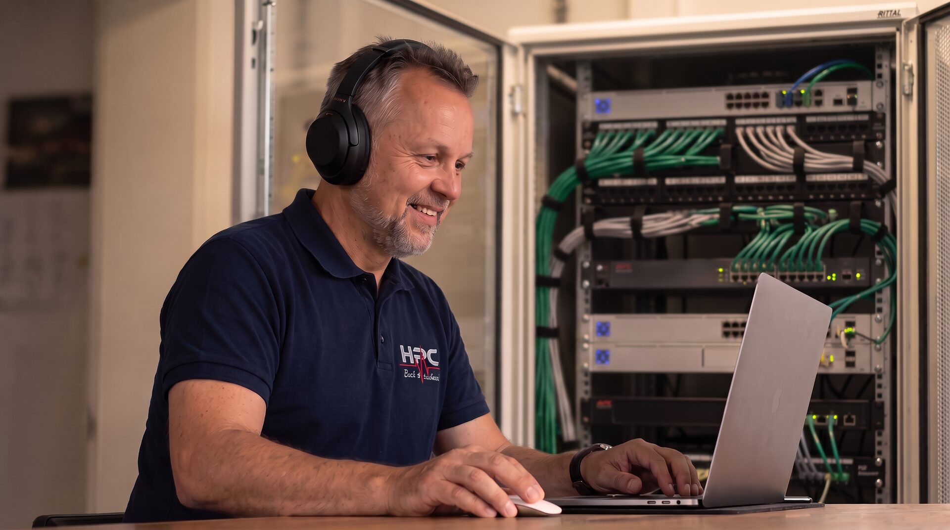Mitarbeiter mit Headset am Notebook vor einem Netzwerkschrank